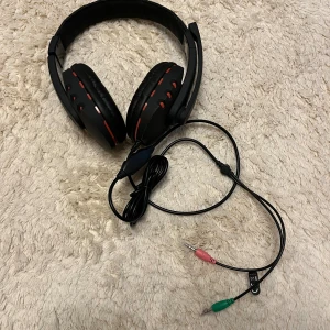Svart gamingheadset DELTACO GAMING - Säljer ett svart gamingheadset från DELTACO GAMING med röda detaljer. Over-ear-modell med mjuka kuddar och justerbar mikrofon. Utrustad med sladd och två 3,5 mm-kontakter, en rosa och en grön, för ljud och mikrofon. Perfekt för gaming och voicechat.