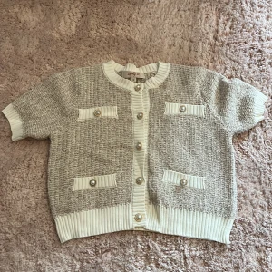 Kortärmad kofta från Derby X - Superfin kortärmad kofta från Derby X i beige och vitt med pärlknappar och dekorativa fickor framtill. Koftan har ribbade kanter och en stickad struktur som ger en lyxig känsla. Perfekt att styla med både jeans och kjol.