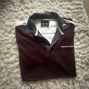 Vinröd pikétröja Peak Performance M - Snygg vinröd pikétröja från Peak Performance med kontrastkrage i mörkgrått och vita detaljer. Broderad logga på bröstet och på kragen. Perfekt för dig som gillar sportig och stilren look.