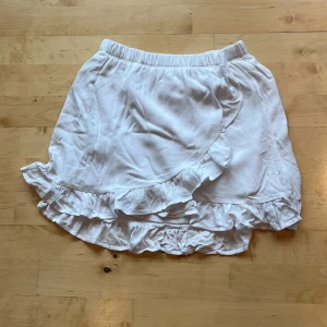 Vit omlottkjol med volang från 157 - Supersöt vit kortkjol från  Lager 157 med omlott-design och volangkant. Kjolen har resår i midjan för en bekväm passform och är gjord i ett lätt och mjukt material, perfekt för varma dagar på stranden. Enkel att matcha med olika toppar för en fräsch look.