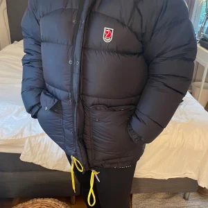Svart dunjacka från Fjällräven XS - Säljer en svart, fin dunjacka från Fjällräven i storlek XS. Jackan har gula snören, hög krage och klassisk Fjällräven-logga på bröstet. Perfekt för kalla dagar med sin pufferstil och stora fickor framtill. Stängs med både dragkedja och knappar. Dragsnöre insida lite defekt. Påverkar inte funktionen. Se bild!