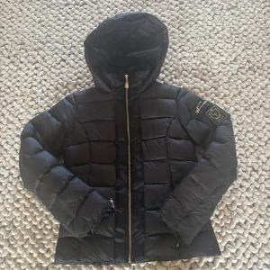 Svart dunjacka från Chiara Forthi - Snygg svart dunjacka från Chiara Forthi Milano med guldiga dragkedjor och emblem på ärmen. Jackan har huva, quiltad design och raka linjer. Perfekt för kalla dagar och ger en clean look.