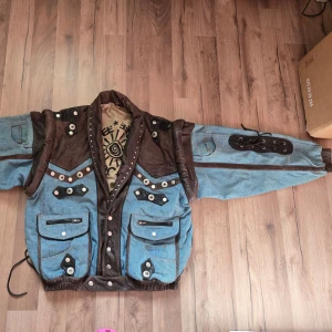 Unik jeans- och skinnjacka med nitar - Säljer en riktigt cool jacka i blått jeanstyg och brunt skinn med massor av detaljer som nitar, knappar och snörning på ärmen. Jackan har flera fickor med dragkedjor och tryckknappar, samt vadderad krage och axelparti. Perfekt för dig som vill sticka ut!