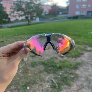 Oakley sportglasögon med spegelglas - Oakley sportglasögon med transparent båge och spegelglas i rosa och orange toner. Sköldformade linser ger bred sikt och skydd, perfekt för cykling eller löpning. Svarta skalmar och näsbrygga för extra komfort och stabilitet.