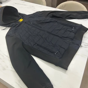 Svart pufferjacka från Parajumpers XL - Svart pufferjacka från Parajumpers i storlek XL. Jackan har quiltad front, släta ärmar, huva med dragsko och flera dragkedjeförsedda fickor. Materialmix av fleece och syntet ger både värme och stil. Snygg gul detalj vid halsen och klassisk logga på ärmen.det står att det är storlek XL men det passar änven som L 