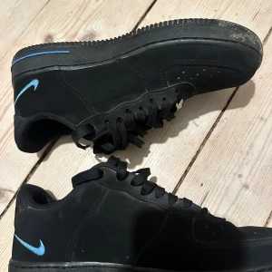 Svarta Nike Air Force 1 sneakers - Svarta Nike Air Force 1 sneakers med klassisk siluett och svart sula. Skorna har svarta skosnören och en blå Nike Swoosh på ena sidan, medan andra sidan har en svart Swoosh. Tillverkade i syntetmaterial med låg profil och rund tå.
