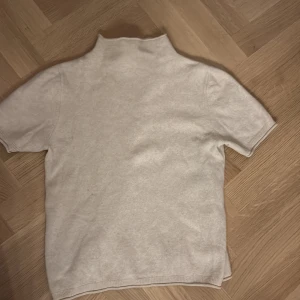 Beige polotröja i ull från RAVS - Beige polotröja från FWSS i mjuk ullmix. Tröjan har korta ärmar, hög krage och en rak passform. Perfekt för lager-på-lager och stilrena outfits. Snygg söm mitt bak och ribbade avslut vid ärmar och nederkant.