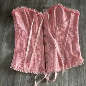 Rosa korsett med spets och snörning - Supersöt rosa korsett med glansigt tyg och brokadmönster. Har volangkant upptill och nedtill samt snörning i ryggen för perfekt passform. Med fina detaljer och metallknäppning framtill. Perfekt för dig som vill sticka ut, även bra nu inför halloween för en look. Endast använt en gång. 