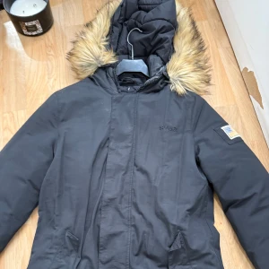 Svart parkajacka med fuskpäls från Svea - Svart parkajacka från Svea med huva och fuskpäls runt kanten. Jackan har broderad amerikansk flagga på ärmen och diskret Svea-logga på bröstet. Praktiska fickor framtill och dragkedja under vindslå. Perfekt för kalla dagar.