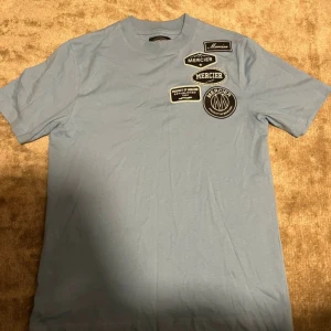 Blå t-shirt från Mercier med patches - Snygg ljusblå t-shirt från Mercier med flera svarta och vita patches på bröstet. Klassisk rund halsringning och korta ärmar. Tillverkad i mjuk bomull för en skön känsla. Perfekt för dig som gillar streetwear och vill sticka ut lite extra.