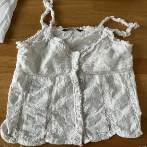 Vit broderad linnetopp från Zara - Supersöt vit linnetopp från Zara med broderade blommor, spetsdetaljer och volangkant. Toppen har knappar framtill och smala, tvinnade axelband. Perfekt för en somrig look med romantiska vibes.