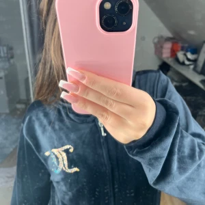 Juicy Couture marinblå velour hoodie - Säljer en marinblå hoodie från Juicy Couture i mjuk velour med glittrande stenar på bröstet och stor Juicy Couture-logga i strass på ryggen. Tröjan har dragkedja framtill, huva och långa ärmar. Perfekt för dig som vill ha den där ikoniska Y2K-looken.