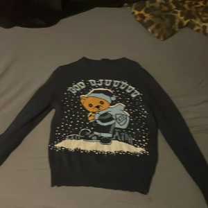 Blå jultröja DIF med björnmotiv - Mörkblå stickad jultröja från Djurgårdens IF med ett stort tryck av en björn i tomteluva och texten 'God Djul'. Tröjan har långa ärmar och ribbade muddar. Perfekt för dig som älskar hockey och vill sticka ut på julfirandet. (Den sitter bättre för dig som har storlek S) pris kan diskuteras. 