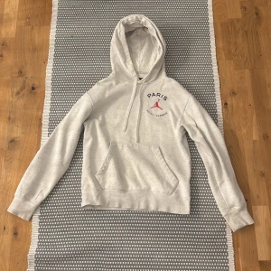 Grå Jordan Paris hoodie XS - Snygg ljusgrå hoodie från Jordan x Paris Saint-Germain med röd logga och text på bröstet samt stor Paris-tryck och siffran 23 på ryggen. Klassisk känguruficka, huva med snörning och mjukt material. Perfekt för en sportig och chill stil.