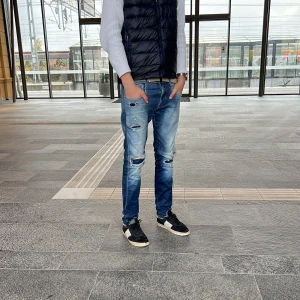 Replay Anbass Hyperflex fast pris - Säljer ett par Replay Anbass Hyperflex jeans i blå tvätt med slitningar och hål på knäna. Jeansen har slim/straight passform, klassisk femficksdesign och stretchigt material för extra komfort. Snyggt slitna detaljer och cool faded effekt. Jeansen är väldigt använda och det kan man se på slitningarna som har gått upp!