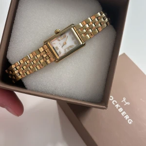 Mockberg guld klocka med länkarmband - Säljer en nästintill oanvänd klocka från mockberg. Urtavlan är vit med romerska siffror och guldfärgade visare. Klockan har en elegant och tidlös design som verkligen sticker ut på handleden. 🤍nypris 1600kr