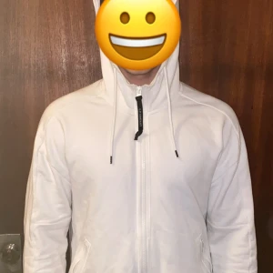 Vit zip-hoodie från CP Company - Säljer en stilren vit hoodie från CP Company med dragkedja framtill och snörning i huvan. Tröjan har långa ärmar, fickor på sidorna och en clean look som passar perfekt till vardag. Hoodien är såklart äkta och köpt på NK, kvitto finns. Den enda som inte gör att den ser ny ut är dragkedjan på ena sidan men något man enkelt kan fixa. Vid fler frågor är det bara att höra av sig😃