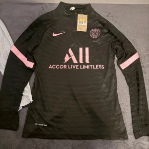 Svart PSG långärmad fotbollströja Nike - Snygg svart långärmad fotbollströja från Nike för Paris Saint-Germain med rosa detaljer och klubbmärke på bröstet. Tröjan har halv dragkedja, Dri-FIT material och diskret mönster i tyget. Perfekt för dig som vill sticka ut på planen.