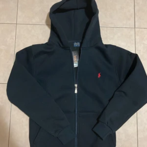 Mörkblå hoodie från Polo Ralph Lauren - Mörkblå hoodie från Polo Ralph Lauren med dragkedja och huva. Klassisk design med röd broderad logga på bröstet. Tillverkad i mjuk bomull, perfekt för en chill och stilren look. Långa ärmar och fickor framtill.