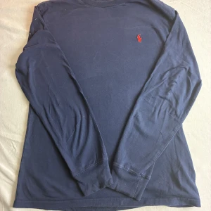 Mörkblå långärmad tröja Polo Ralph Lauren - Klassisk mörkblå långärmad tröja från Polo Ralph Lauren med röd broderad logga på bröstet. Tillverkad i mjuk bomull, perfekt för en stilren och avslappnad look. Rund halsringning och normal passform. Storlek 