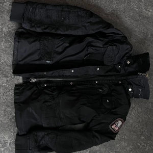 Svart vinterjacka Parajumpers med päls - Säljer en svart vinterjacka från Parajumpers med pälsfodrad insida och huva. Jackan har flera fickor, patch på ärmen, quiltade detaljer och en gul rem vid kragen. Stängs med dragkedja och knappar. Perfekt för kalla dagar.