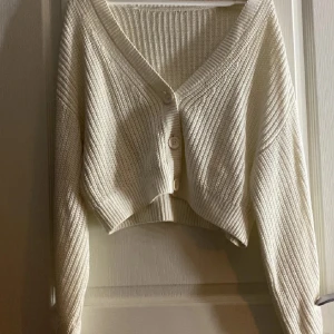Beige croppad kofta med knappar - Superfin beige croppad kofta med v-ringning och stora knappar framtill. Stickad i ett mjukt material och har långa ärmar. Perfekt att slänga över en topp för en chill look. Storleks lappen är avklippt(vet ej vilken strl)