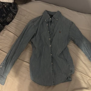 Blå jeansskjorta från Ralph Lauren - Snygg blå jeansskjorta från Ralph Lauren i slim fit-modell. Skjortan har klassisk krage med knappar, långa ärmar och knäppning framtill. Ikonisk broderad logga på bröstet. Perfekt för en avslappnad men stilren look.