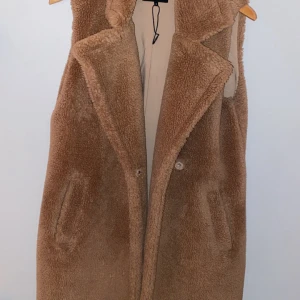 Beige teddyväst från ONLY - Mysig beige teddyväst från ONLY med bred krage och två fickor framtill. Västen är lång och har en klassisk rak passform, perfekt att slänga över en hoodie eller tröja för extra värme. Stängs med knappar framtill och har ett mjukt foder.
