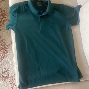 Mörkgrön pikétröja från Polo Ralph Lauren - Snygg mörkgrön pikétröja från Polo Ralph Lauren med klassisk krage och tre knappar framtill. Tröjan har korta ärmar och den ikoniska broderade loggan på bröstet. Perfekt för dig som gillar stilrena och sportiga plagg.