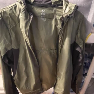 Grön vindjacka med huva från SOC - Snygg grön vindjacka från SOC med svarta detaljer på sidorna och ärmarna. Jackan har huva, hel dragkedja framtill och är tillverkad i lätt polyester. Perfekt för blåsiga dagar och enkel att slänga på sig när du är på språng.