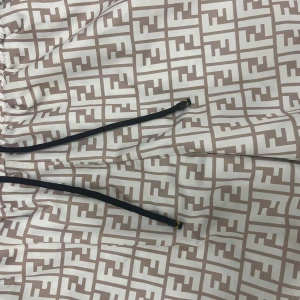 Beige och vita Fendi shorts med logomönster - Snygga shorts från Fendi i beige och vitt med ikoniskt FF-logomönster över hela tyget. De har elastisk midja med svart snörning och innerfoder i mesh. Perfekta för dig som vill sticka ut med en lyxig streetstyle-look.