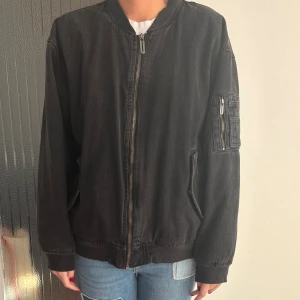 Svart bomberjacka i denim från New Yorker - Snygg svart jeans bomberjacka i denim från FB Sister. Jackan har klassisk bombermodell med ribbade muddar, dragkedja framtill och cool ficka med dragkedja på ärmen. Perfekt oversized fit för en avslappnad streetstil. Den är varm. Jag har storlek S. 