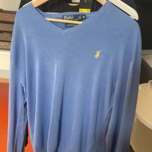 Blå v-ringad tröja från Polo Ralph Lauren - Snygg blå v-ringad tröja från Polo Ralph Lauren i 100% pima bomull. Klassisk design med gul broderad logga på bröstet och ribbade muddar. Perfekt för dig som gillar stilrena och tidlösa plagg.