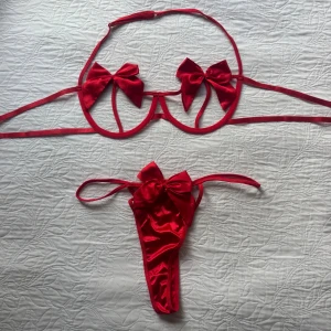 Röda underkläder med rosetter och kedjedetaljer - Superfin sexiga underkläder med stora rosetter på både överdel och underdel. Överdelen har öppna kupor med två satinskimrande rosetter och smala band. Underdelen är stringmodell med en matchande rosett framtill. Tillbehör med rött midjeband, hjärtformad detalj och silverfärgade kedjor ingår. Använd en gång/provade. Storlek XL, men förminskad som L.