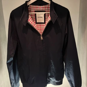 Svart vindjacka från Ben Sherman M - Stilren baby blå harrington jacka från Ben Sherman i storlek M. Jackan har dragkedja framtill, klassisk krage med två knappar och ett rutigt innerfoder i rött, vitt och grått. Perfekt för dig som gillar en clean look med snygga detaljer. 