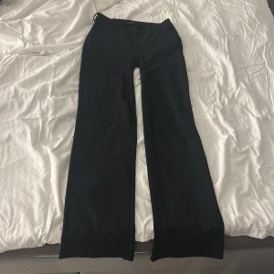 Svarta vida byxor från Vero Moda - Svarta kostymbyxor från Vero Moda med vida ben och klassisk passform. Byxorna har hällor för bälte och stängs med dragkedja och knapp. Perfekta för en clean och stilren look. Materialet känns mjukt och följsamt.