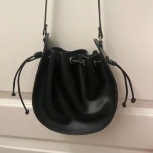 Svart bucket bag med snörning - Snygg svart bucket bag i skinnimitation med justerbar axelrem och cool snörning upptill. Väskan har rundad form och metallöglor för snörningen, perfekt för dig som vill ha en stilren och trendig accessoar.