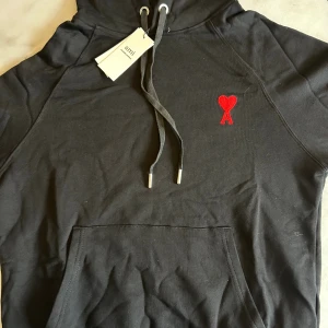 Svart hoodie från Ami med rött hjärta - Svart hoodie från Ami Alexandre Mattiussi med klassisk huva och snörning. På bröstet finns ett broderat rött hjärta med bokstaven A. Hoodien har känguruficka och är tillverkad i mjuk bomull, perfekt för en chill och stilren look.