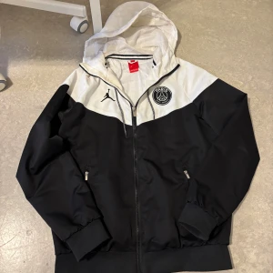 PSG x Jordan svartvit vindjacka - Svart och vit vindjacka från Jordan i samarbete med Paris Saint-Germain. Jackan har huva, dragkedja framtill, två fickor med dragkedja och PSG-logga på bröstet. Tillverkad i lätt polyester med meshfoder, perfekt för blåsiga dagar. Pris kan diskuteras!