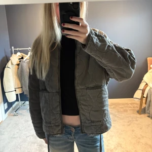 Grå quiltad jacka från Zara XS - Quiltad jacka från Zara i grått med vågigt mönster i tyget. Jackan har en loose fit, två stora fickor framtill och hög krage. Perfekt för lager-på-lager och chill stil. Stängs utan dragkedja eller knappar.