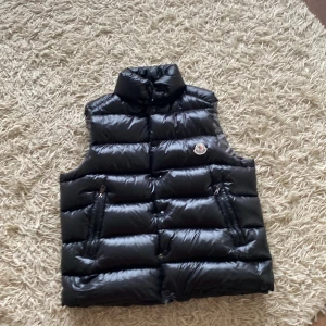 Svart dunväst från Moncler - Säljer en svart, glansig dunväst från Moncler med klassisk quiltad design och hög krage. Västen har tryckknappar framtill och ikonisk Moncler-logga på bröstet. Insidan har ett coolt serietidningsmönster med tvättråd. Perfekt för lager-på-lager-stil.