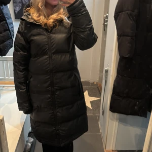 Svart lång pufferjacka med huva - Säljer en svart lång pufferjacka med huva och gul insida. Jackan har dragkedja hela vägen, två fickor och ribbade muddar vid ärmsluten. Den är vattentät och är vaxad, perfekt inför vintern.