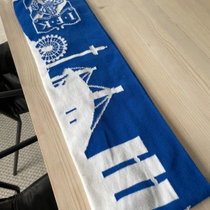 IFK Göteborg halsduk blåvit - Blåvit halsduk med IFK Göteborgs klubbmärke och motiv från Göteborg, inklusive Lisebergshjulet och Ullevi. Halsduken har vita fransar i ändarna och är perfekt för att visa ditt stöd på läktaren. Tjockt stickat material som håller dig varm under matchen.