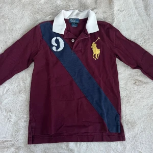 Bordeaux pikétröja från Ralph Lauren - Snygg bordeaux pikétröja från Ralph Lauren med vit krage, lång ärm och klassisk gul broderad logga. Tröjan har en marinblå diagonal rand och vit siffra på bröstet. Perfekt för dig som gillar sportig och preppy stil.
