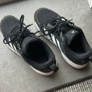 Svarta Adidas sneakers strl 36 - Säljer ett par svarta Adidas sneakers som är inneskor med vita detaljer och klassiska ränder på sidorna. Skorna har snörning, platt sula i gummi och ovandel i textil. Perfekta för dig som gillar sportig stil och vill ha bekväma skor till vardagen.