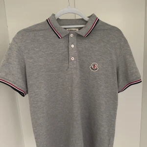 Grå pikétröja från Moncler (S) - Snygg grå pikétröja från Moncler i klassisk modell med krage och knappar. Tröjan har Moncler-logga på bröstet och detaljer i rött, vitt och blått på krage och ärmslut. Tillverkad i mjuk bomull och perfekt för en clean och sportig stil. OBS: den har en liten fläck längre ner i ryggen, som man kan få bort med medel o tvätt.