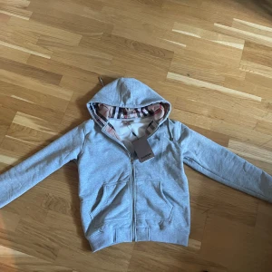 Grå Burberry hoodie med klassiskt foder - Snygg grå hoodie från Burberry med dragkedja och huva. Insidan är fodrad med det ikoniska rutiga Burberry-mönstret i beige, svart och rött. Tröjan har två fickor framtill och ribbade muddar. Perfekt för dig som vill ha en stilren och lyxig vardagslook.