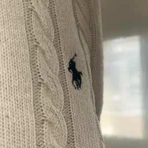 Vit kabelstickad tröja Polo Ralph Lauren - Vit kabelstickad tröja från Polo Ralph Lauren med klassisk rund halsringning och broderad mörkblå logga på bröstet. Tröjan har ribbade muddar och är långärmad, perfekt för lager-på-lager under höst och vinter.
