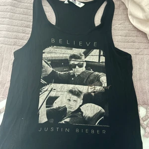 Justin Bieber linne Believe svart - Svart linne med tryck från Justin Biebers Believe-era. Framsidan har ett stort svartvitt foto av Justin Bieber i bil och texten 'BELIEVE' samt 'JUSTIN BIEBER'. Klassisk ärmlös modell med brottarrygg. 