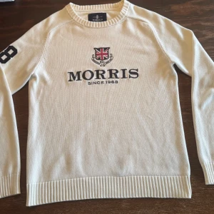 Vit stickad tröja från Morris - Snygg vit stickad tröja från Morris med broderad logga och brittisk flagga på bröstet. Tröjan har ribbade muddar, rund hals och svarta siffror på ena ärmen. Perfekt för dig som gillar klassisk och stilren design.
